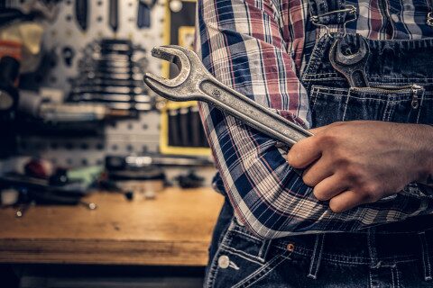 A mechanic in a checked shirt and dungarees holds a large wrench in front of him.